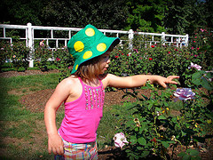 little girl wearing a green bucket hat with yellow polka dots in a garden pointing at flowers 