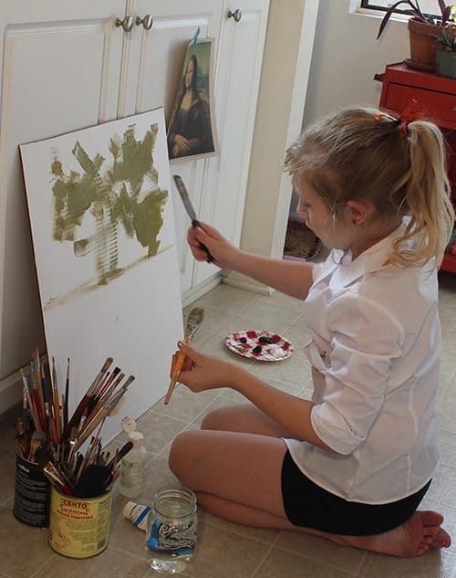 a child sitting on the floor next to cans of paint brushes,  painting on cardboard 