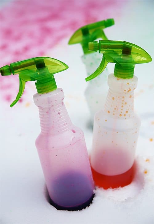close-up photo of three spray bottles filled with purple, pink, and green food dye placed into the snow outdoors