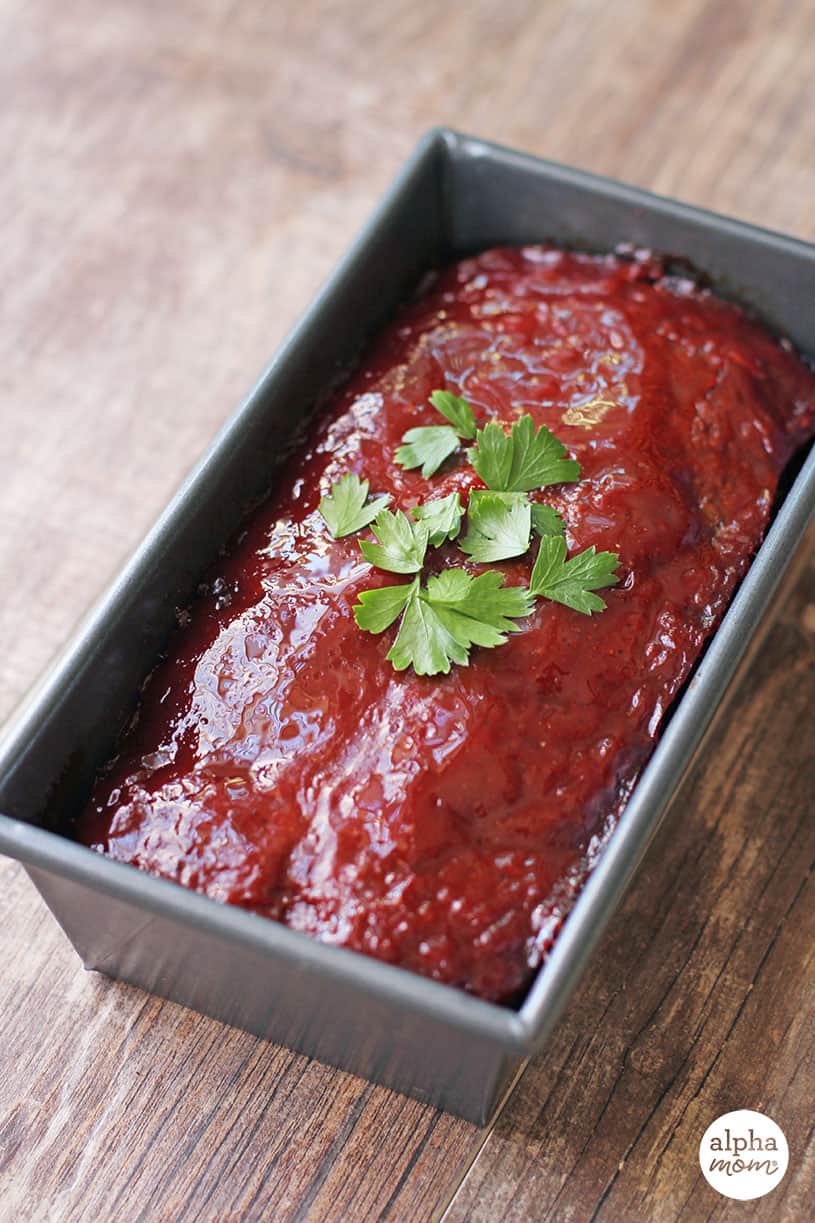 Vertical photo side view of cooked meatloaf in a pan with ketchup glaze and parsley garnish