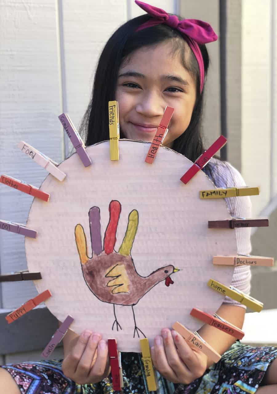 Smiling young girl holding cardboard with handprint turkey and clothespins with gratitude mantras 