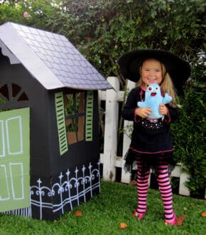 Smiling little girl dressed in witch costume holding a ghost stuffie next to a black life-sized haunted house made from cardboard boxes with white roof and green door
