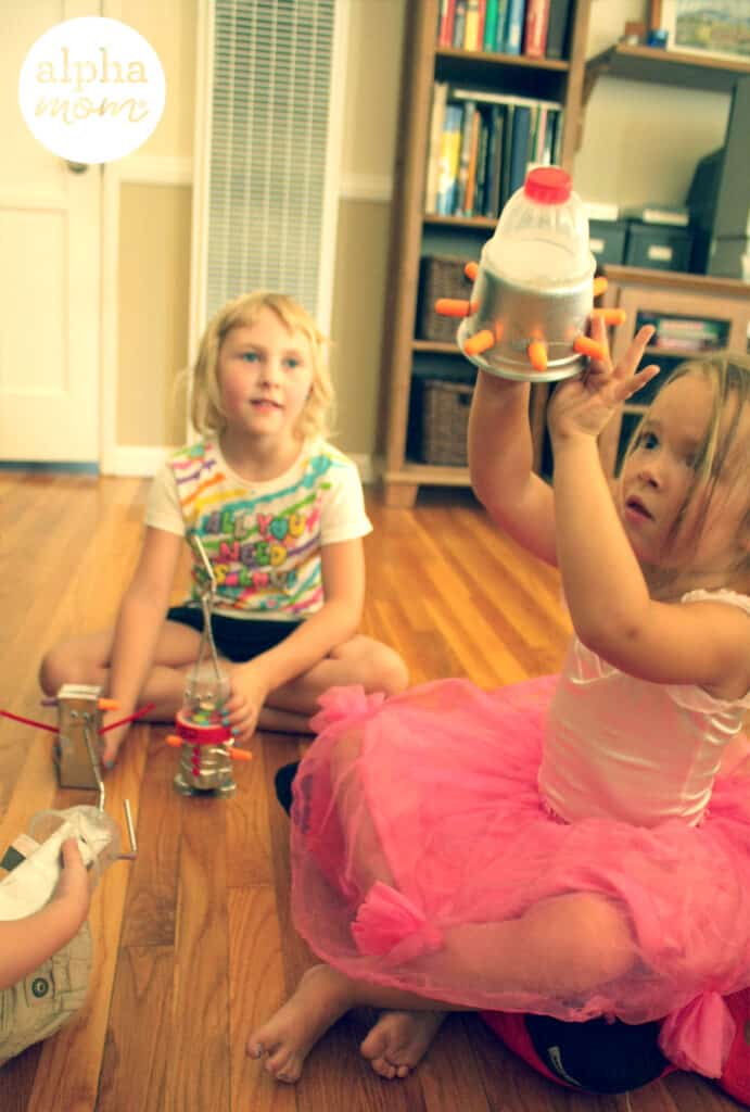 photo of two girls sitting on the floor playing and one is wearing a pink tutu and holding up a handmade toy and the other is looking at her