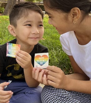young Asian boy and girl looking at each other and sitting on grass while holding up single Rainbow Valentine Card