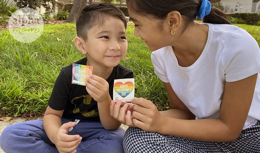young Asian boy and girl looking at each other and sitting on grass while holding up single Rainbow Valentine Card