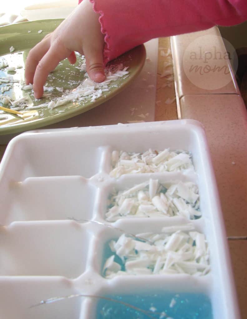close-up of child's hand sprinkling shaved white soap from green plate into ice cube tray to make handmade Snowglobe soap
