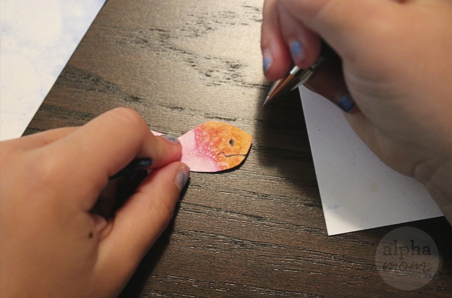 close-up of hand drawing eyes and mouth with pen onto pink and orange paper fish art 