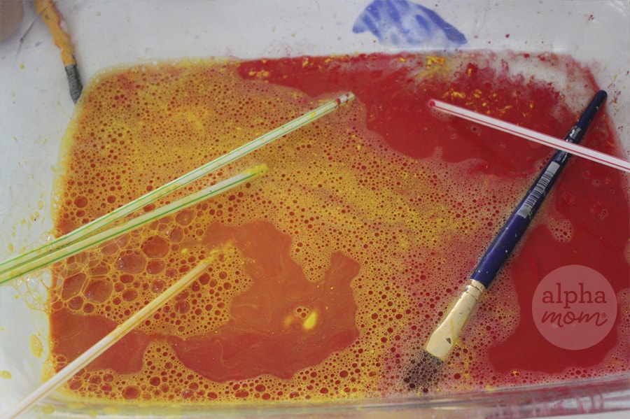 soapy red paint water in glass dishpan with plastic straws and paint brush strewn about
