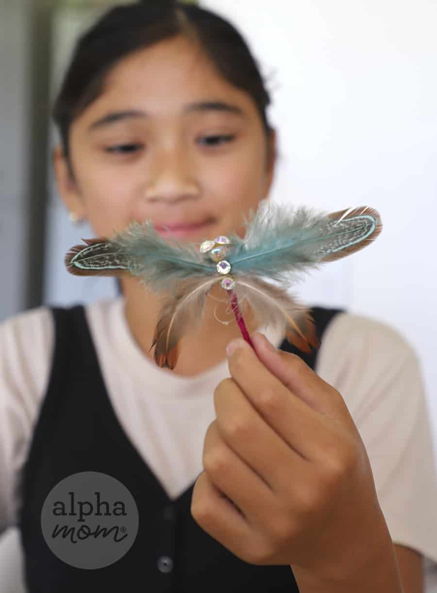Girl with dark hair pulled back wearing white shirt and black vest holding up and inspecting a turquoise feather dragonfly hairpin in front of her