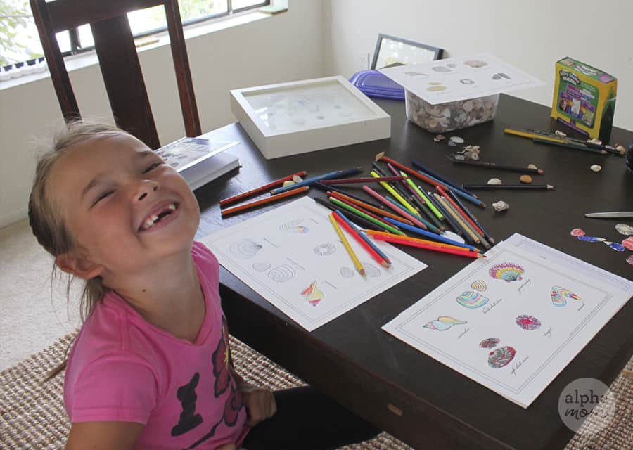laughing young girl in pink t-shirt tossing her back and looking at the camera at activity table coloring in sea shell printable with lots of coloring pencils in front of her and sea shells on the table