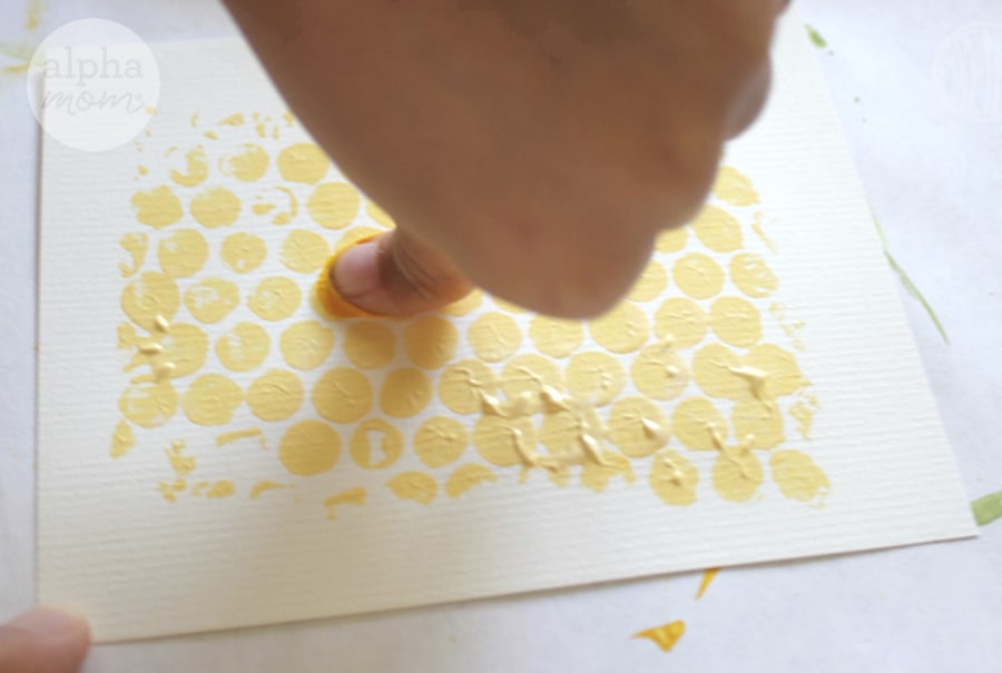 close-up of girl's thumb pressing darker yellow paint and stamping thumbprints on cardstock painted with yellow bubblewrap print to make Rosh Hashanah Honey Bee Cards