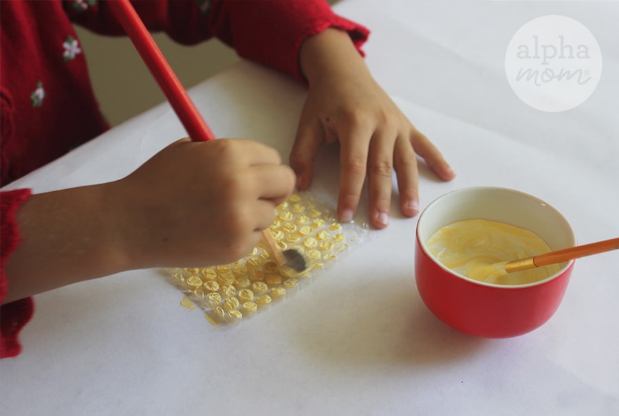 Action photo of close-up of hands of girl in red shirt painting bubble wrap with yellow paint at white desk with red bowl filled with paint in front of her